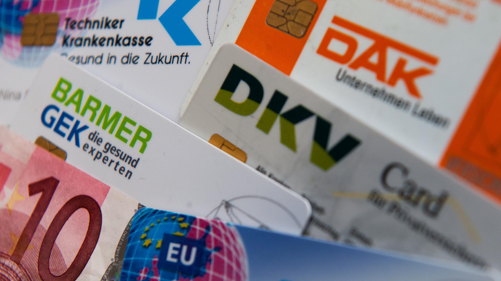 Zu sehen sind viele verschiedene Krankenkassenkarten bunt ineinander gewürfelt. Dazwischen klemmt versteckt ein zehn Euro-Schein.