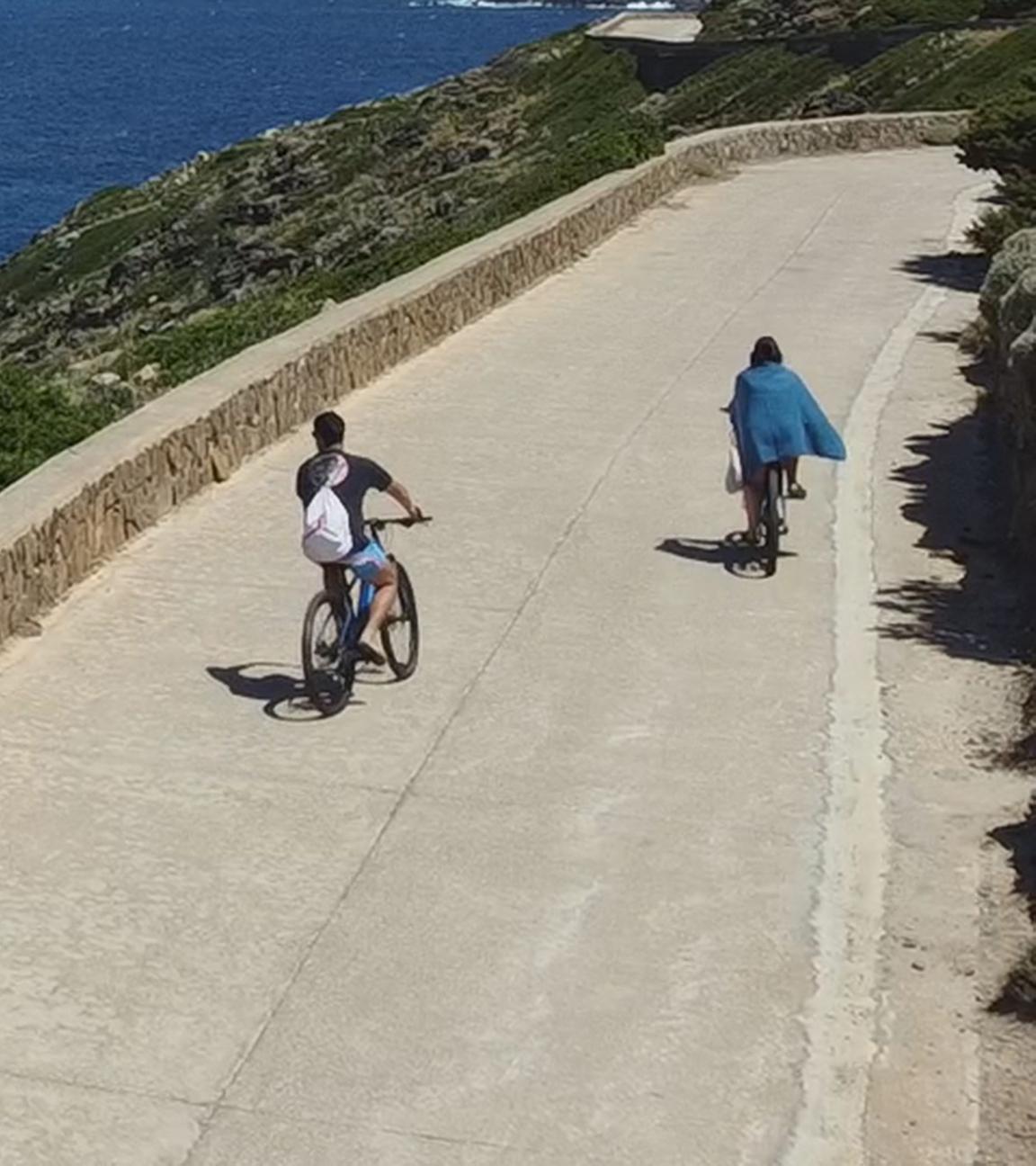 Landschaftspanorama: Oben an der Küste der Insel Asinara entlang verläuft eine Straße, auf der sich zwei Fahrradfahrer befinden. Dahinter erstreckt sich das tiefblaue Meer unter einem blauen Himmel bis zum Horizont.