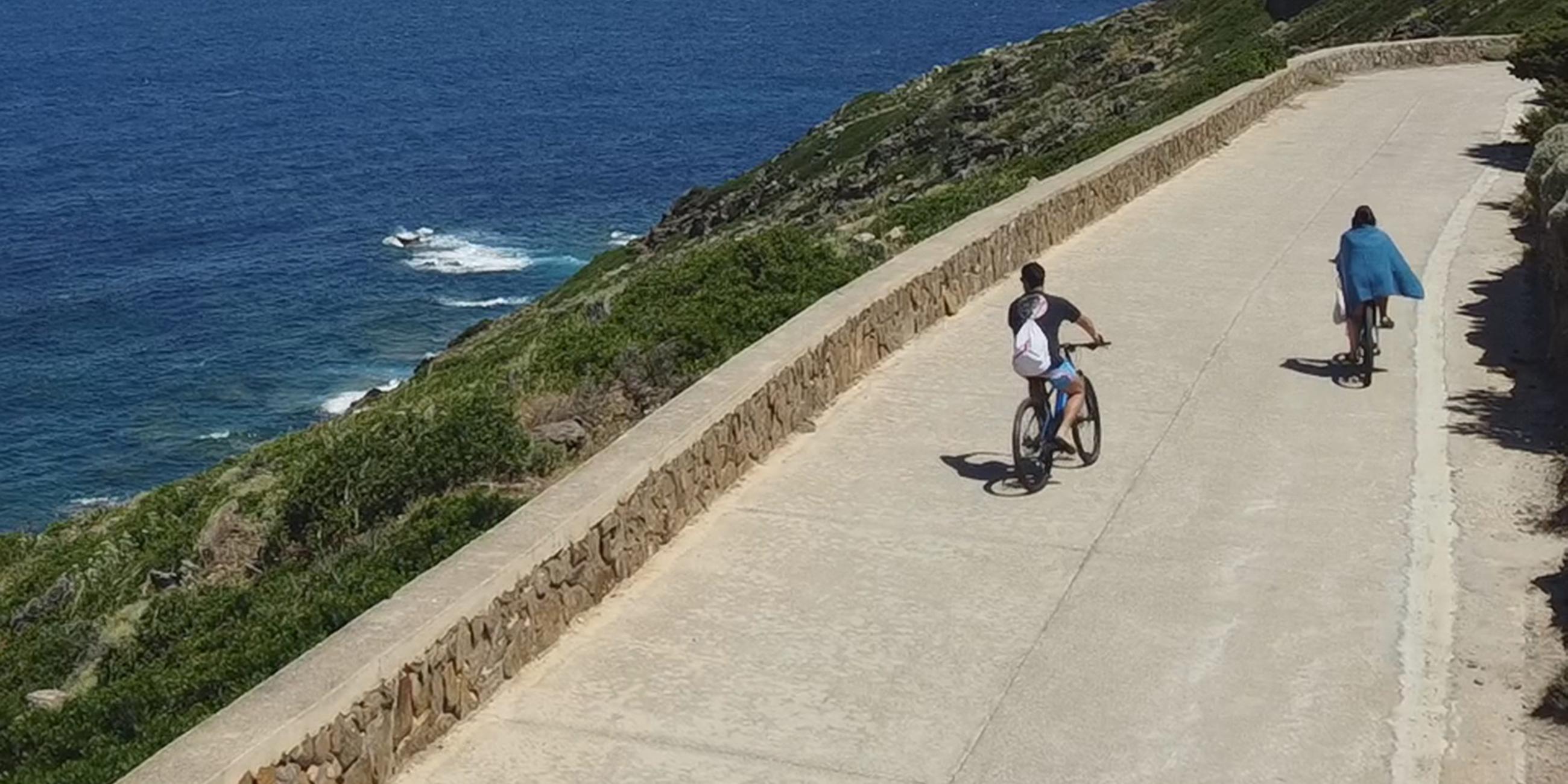 Landschaftspanorama: Oben an der Küste der Insel Asinara entlang verläuft eine Straße, auf der sich zwei Fahrradfahrer befinden. Dahinter erstreckt sich das tiefblaue Meer unter einem blauen Himmel bis zum Horizont.