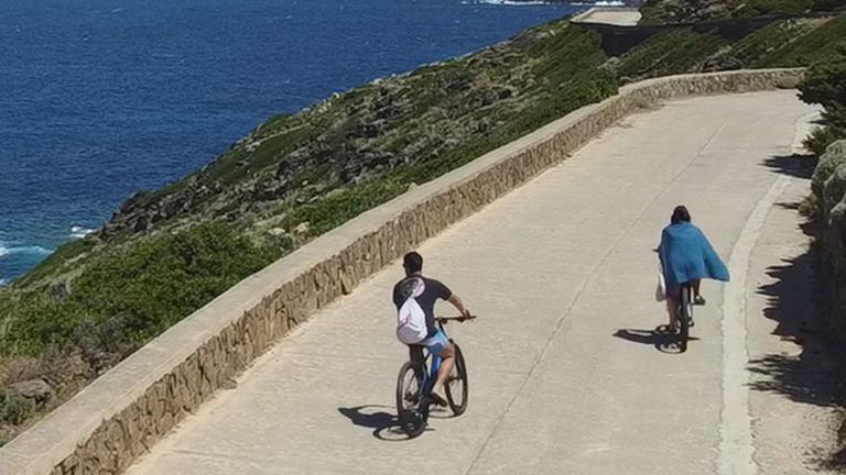 Landschaftspanorama: Oben an der Küste der Insel Asinara entlang verläuft eine Straße, auf der sich zwei Fahrradfahrer befinden. Dahinter erstreckt sich das tiefblaue Meer unter einem blauen Himmel bis zum Horizont.