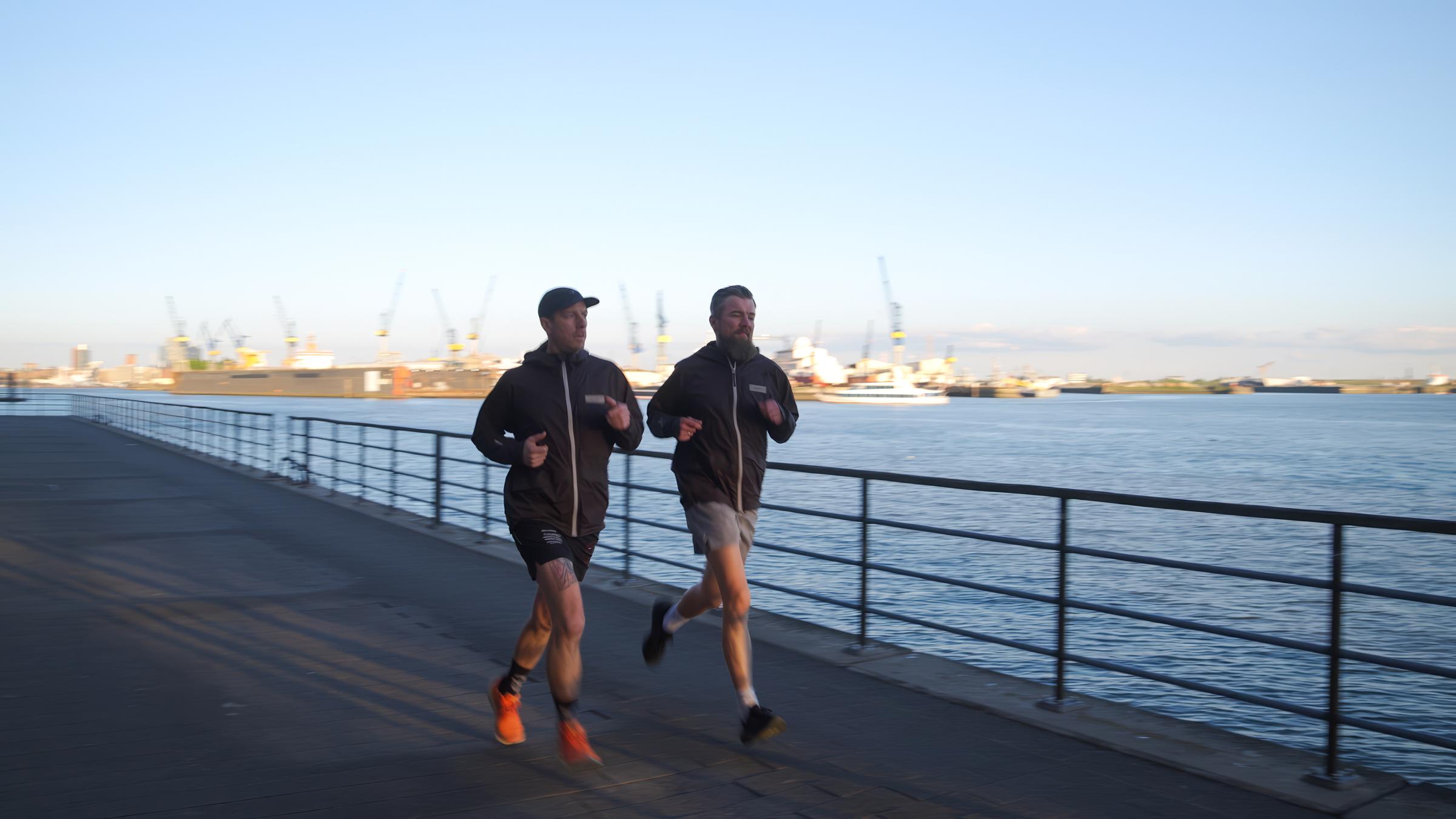 Zwei Männer joggen an einem befestigten Ufer entlang. Im Hintergrund liegen Schiffe im Hafen, Kräne ragen empor.