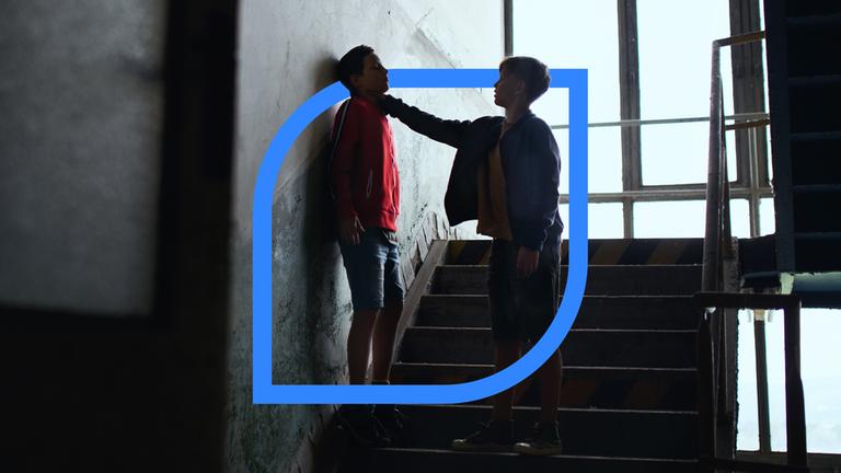 Zwei Jugendliche stehen in einem Treppenhaus. Der eine mit dem Rücken zur Wand, während der andere ihn an den Hals fässt. 