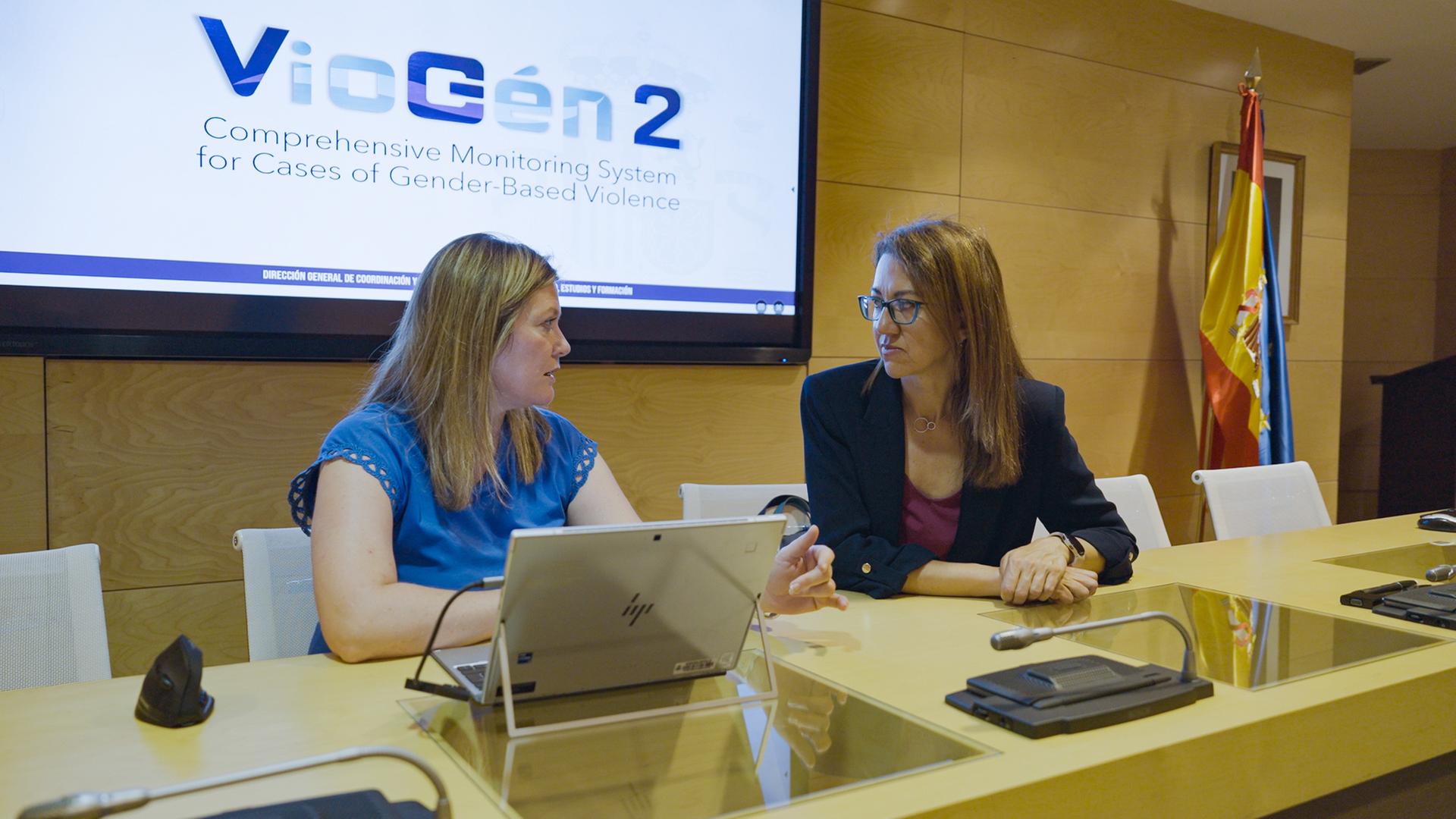 Belén Crego Sánchez von der spanischen Polizei sitzt mit einer Kollegin an einem Konferenztisch. Sie schauen sich an und unterhalten sich. Hinter ihnen auf einem Display steht "VioGén2".