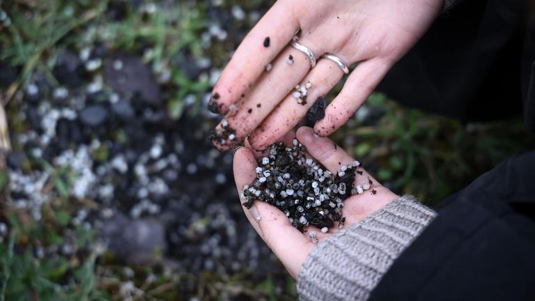 Eine Person sammelt Plastikpellets auf dem Boden in der Nähe eines Industriegebiets.