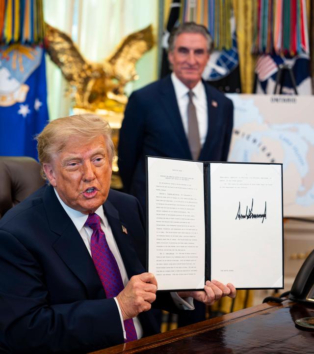 Donald Trump sitzt an seinem Schreibtisch im Oval Office und hält ein unterschriebenes Blatt Papier hoch.