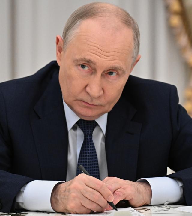 Russlands Präsident Wladimir Putin lehnt sich über den Schreibtisch und blickt in Richtung Kamera.