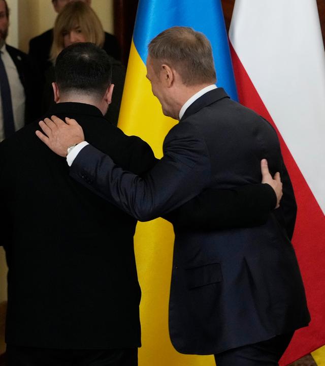 Selenskyj und Tusk halten sich an den Armen und drehn sich von der Kamera weg. Im Hintergrund stehen ukrainische und polnische Flaggen.