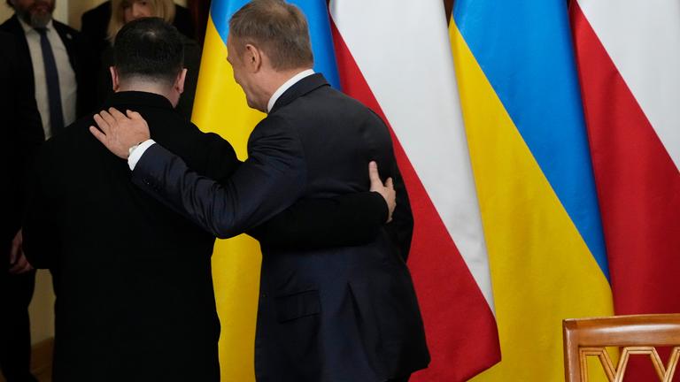Selenskyj und Tusk halten sich an den Armen und drehn sich von der Kamera weg. Im Hintergrund stehen ukrainische und polnische Flaggen.