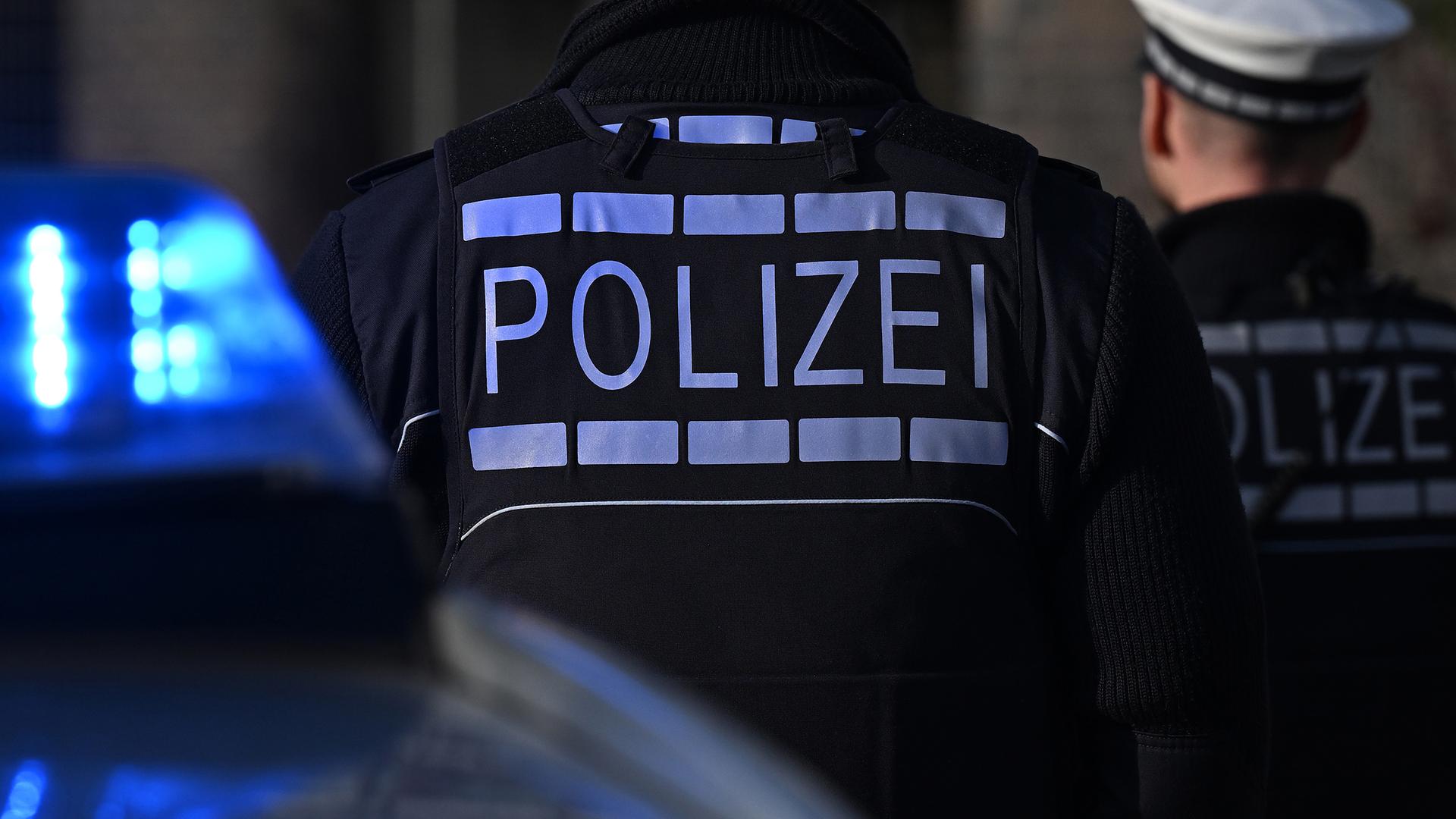 Das Logo der Polizei auf der Dienstkleidung von Polizisten die vor einem Blaulicht stehen