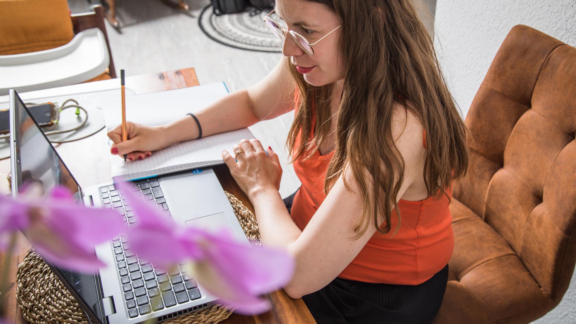 Eine Frau sitzt auf einem Stuhl an einem Tisch vor einem Laptop. Neben dem Laptop schreibt sie etwas auf ein Papier.
