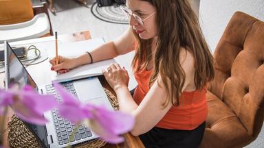 Eine Frau sitzt auf einem Stuhl an einem Tisch vor einem Laptop. Neben dem Laptop schreibt sie etwas auf ein Papier.