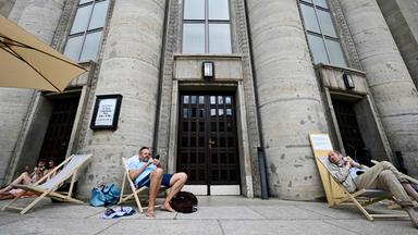 Am Tag der Eröffnung, dem 07.08.2026, sitzen Schwimmer auf Liegestühlen vor dem sogenannten „Volksbad“, das vor der Volksbühne am Rosa-Luxemburg-Platz in Berlin eingerichtet wurde.