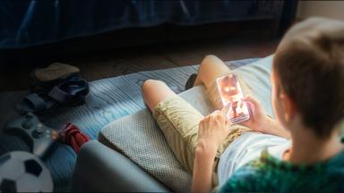 Ein Junge sitzt im Anschnitt mit dem Rücken zur Kamera auf seinem Bett und schaut auf sein Smartphone. Auf dem Display ist undeutlich eine Frau in Unterwäsche abgebildet.