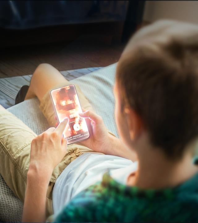 Ein Junge sitzt im Anschnitt mit dem Rücken zur Kamera auf seinem Bett und schaut auf sein Smartphone. Auf dem Display ist undeutlich eine Frau in Unterwäsche abgebildet.