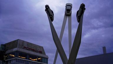 Drei Autos auf hohen Säulen neben einem dem Gebäude des Autoherstellers Porsche in Stuttgart bei Abendstimmung.