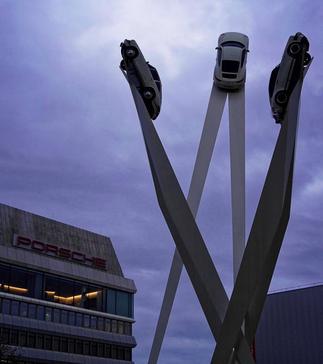 Drei Autos auf hohen Säulen neben einem dem Gebäude des Autoherstellers Porsche in Stuttgart bei Abendstimmung.