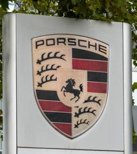 Das Porsche aussen am Gebaeude, Das Porsche-Logo ist ein goldfarbenes Wappen mit rotem und schwarzem Streifenmuster, Hirschgeweihen, einem springenden schwarzen Pferd und der Aufschrift Stuttgart, gekroent vom Schriftzug Porsche.