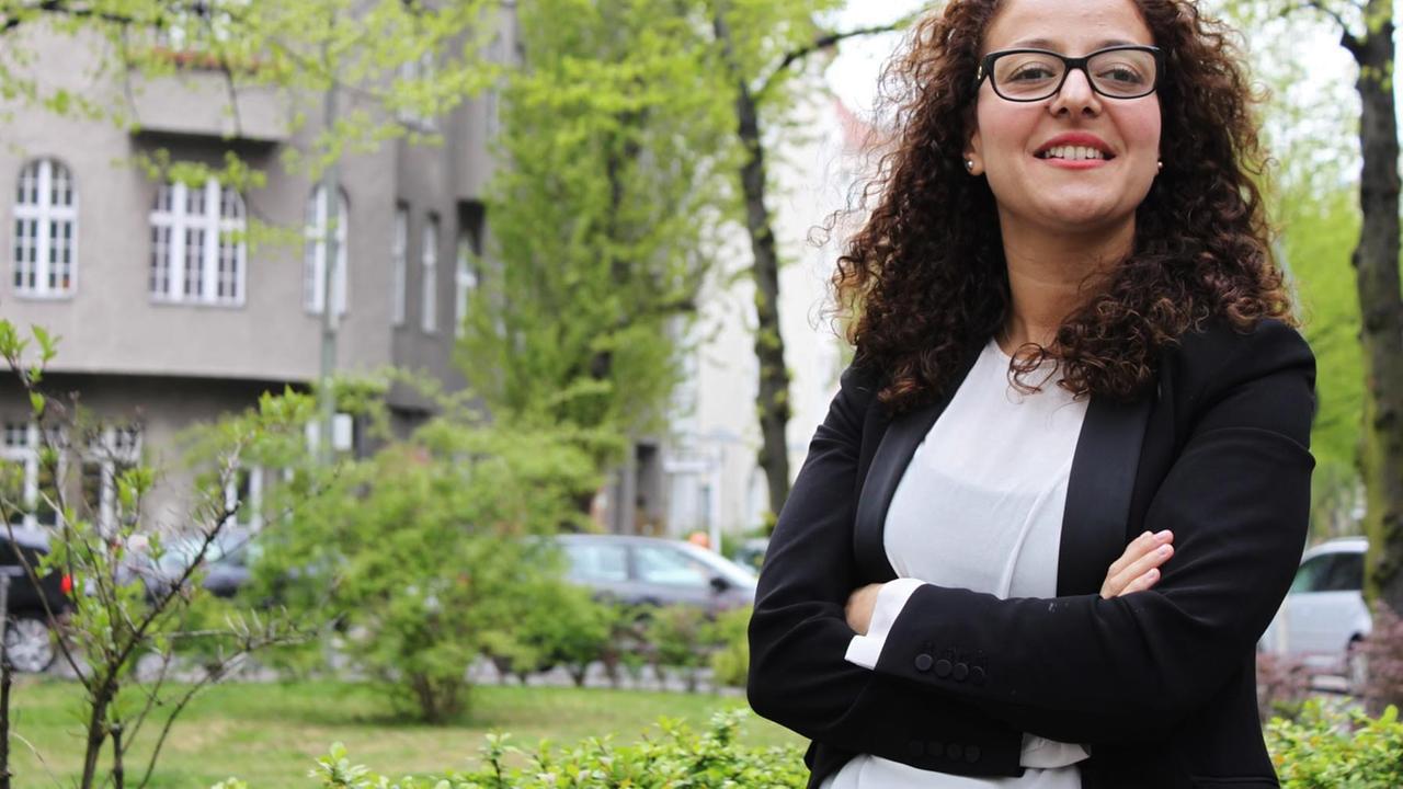 Eine Frau mit dunklen Haaren und Brille steht mit verschränkten Armen in einem Park. Sie lächelt freundlich.