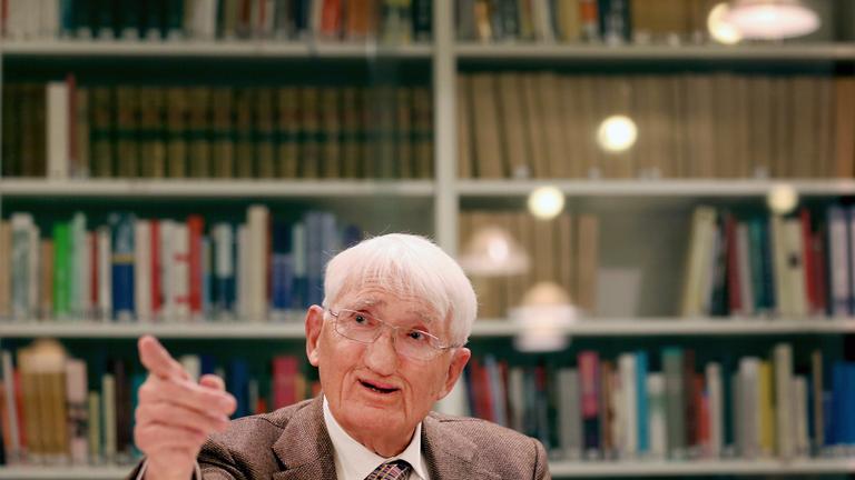 Jürgen Habermas sitzt vor einem Bücherregal und zeigt mit dem Finger an der Kamera vorbei.