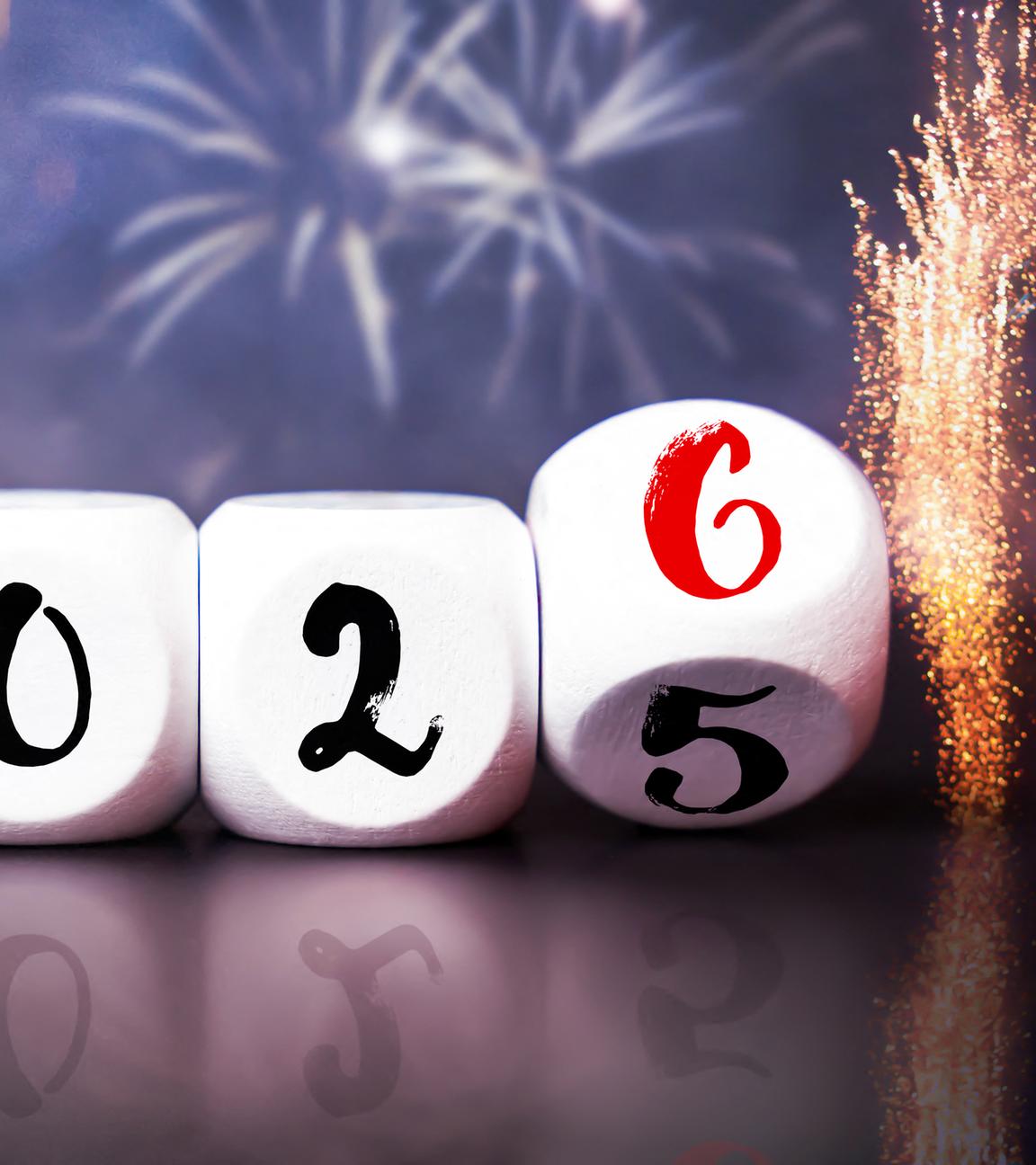 Würfel mit der Aufschrift "2025" bzw. "2026" liegen auf einem Tisch. Im Hintergrund ist ein Feuerwerk zu erkennen.