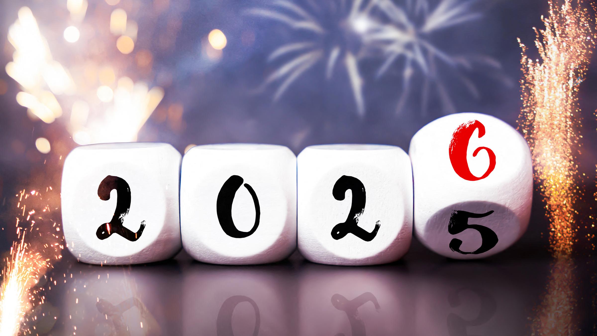 Würfel mit der Aufschrift "2025" bzw. "2026" liegen auf einem Tisch. Im Hintergrund ist ein Feuerwerk zu erkennen.
