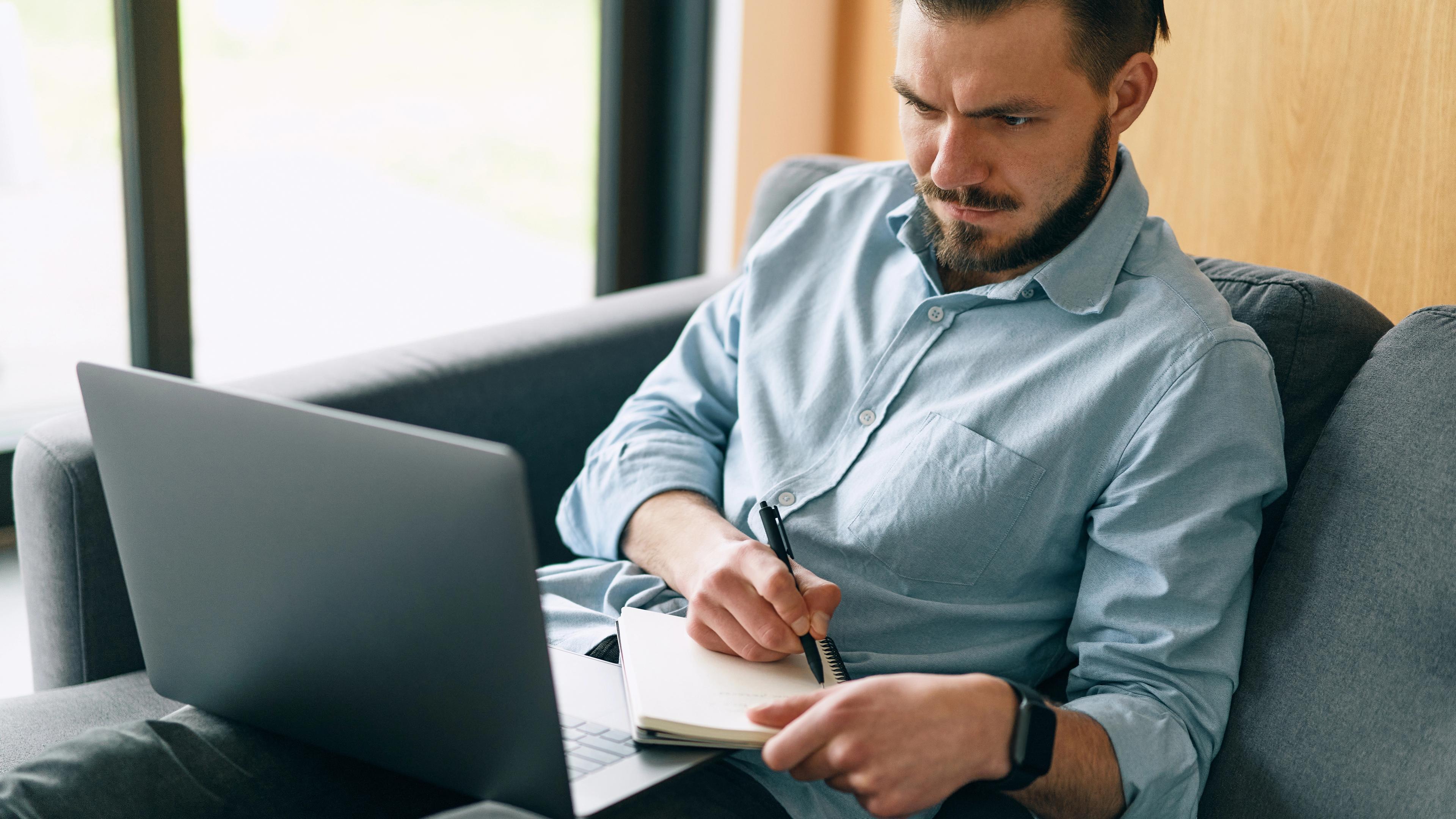 Ein Mann sitzt mit verbissenem Gesicht vor dem Laptop und schreibt eifrig auf einen Block.  