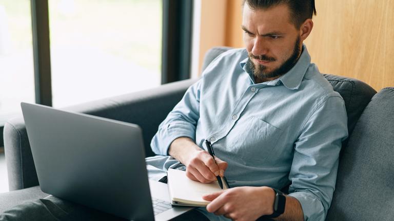 Ein Mann sitzt mit verbissenem Gesicht vor dem Laptop und schreibt eifrig auf einen Block.  