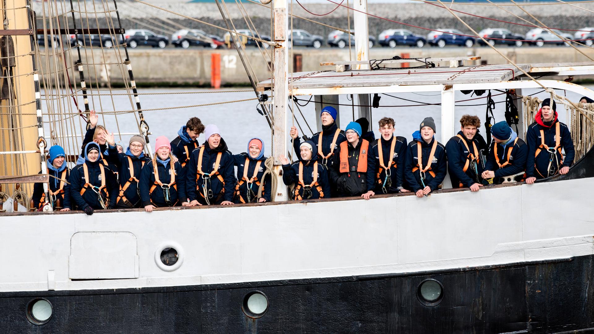 Schülerinnen und Schüler begrüßen bei der Einfahrt in den Hafen von Bord des Schulschiffs "Pelican of London" ihre an Land wartenden Angehörigen am 10.04.2021, Niedersachsen, Emden.