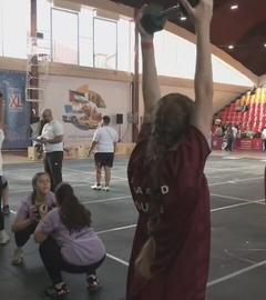 Zwei Mädchen in einer Sporthalle mit Tribüne recken jeweils eine Hantel in die Luft, im Hintergrund sind weitere Menschen sportlich aktiv.