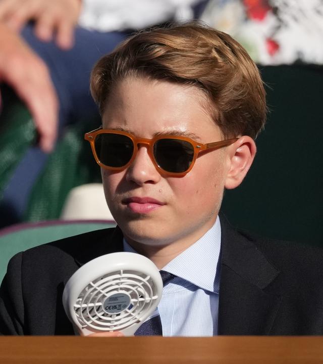 Ein Junge trägt eine Sonnenbrille, einen Anzug und erfrischt sich mit einem Ventilator.