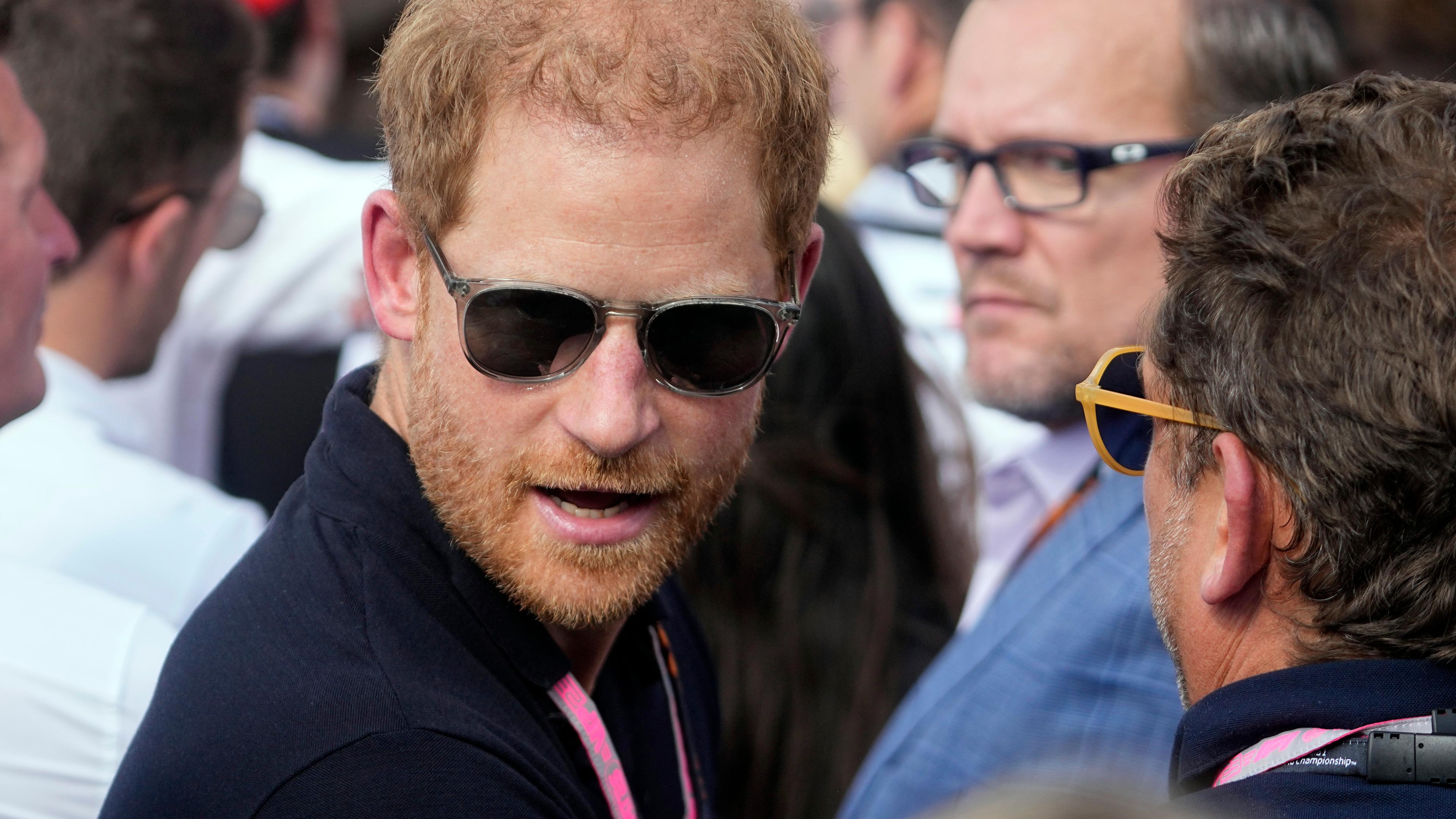 Prinz Harry trägt eine Sonnenbrille und steht zwischen mehrerem Menschen.