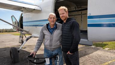 Auf dem Bild sind zu sehen: Ed Marshall und Prinz Harry posieren mit einem Rollator auf dem Rollfeld neben einem Flugzeug.