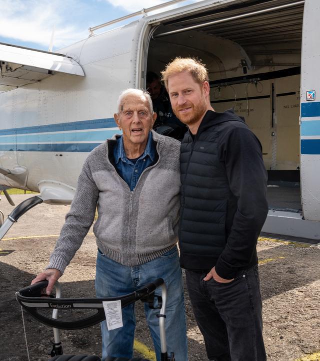 Auf dem Bild sind zu sehen: Ed Marshall und Prinz Harry posieren mit einem Rollator auf dem Rollfeld neben einem Flugzeug.