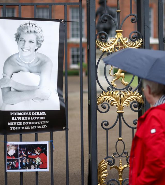 Eine Person mit Regenschirm schaut Bild Prinzessin Dianas am Kensington Palace an