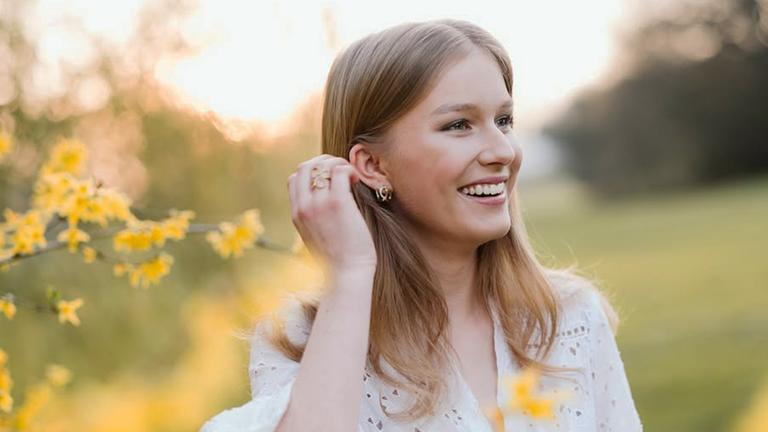 Prinzessin Eléonore ist zu sehen, sie befindet sich in der Natur und im Vordergrund sind einzelne Blumen sichtbar.