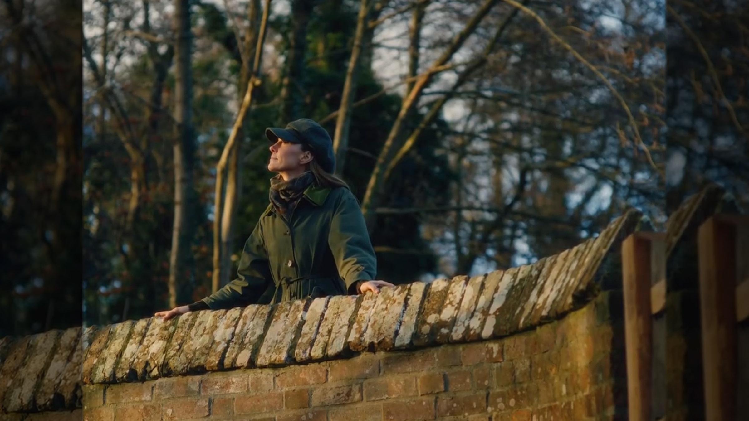 Prinzessin Kate steht in einer nachdenklichen Pose in einem Wald und stützt sich dabei an einer Mauer aus Ziegelsteinen ab.