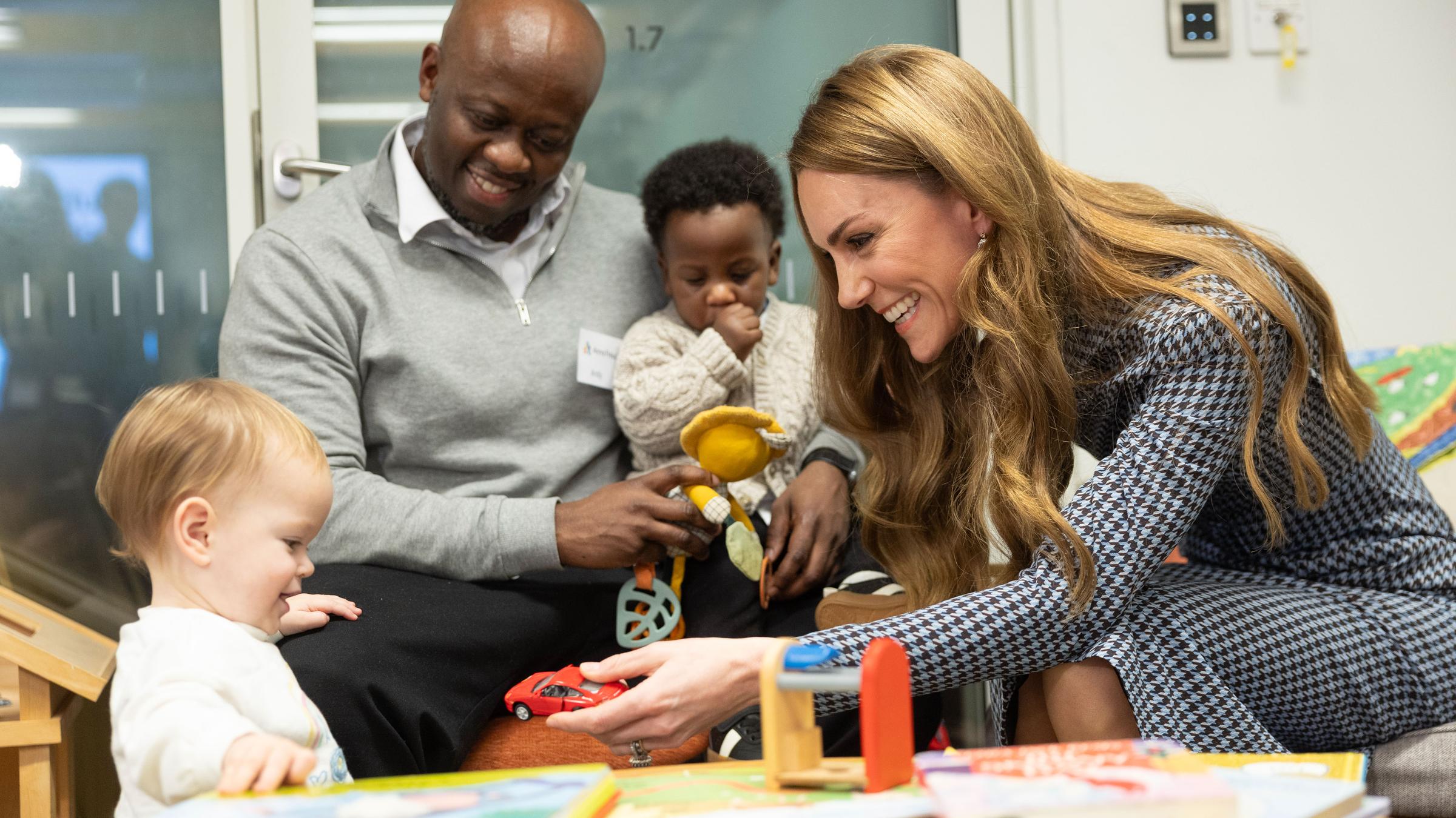 Prinzessin Kate spielt mit einem Kleinkind. Im Hintergrund sitzt ein Mann mit einem weiteren Kleinkind.