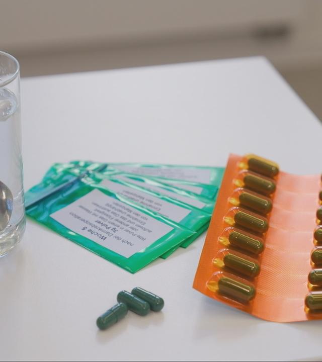 Auf einem Tisch liegen verschiedene Tabletten. Daneben steht ein Glas Wasser mit einem kleinen Löffel drin.