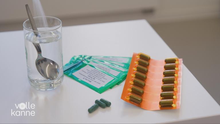 Auf einem Tisch liegen verschiedene Tabletten. Daneben steht ein Glas Wasser mit einem kleinen Löffel drin.