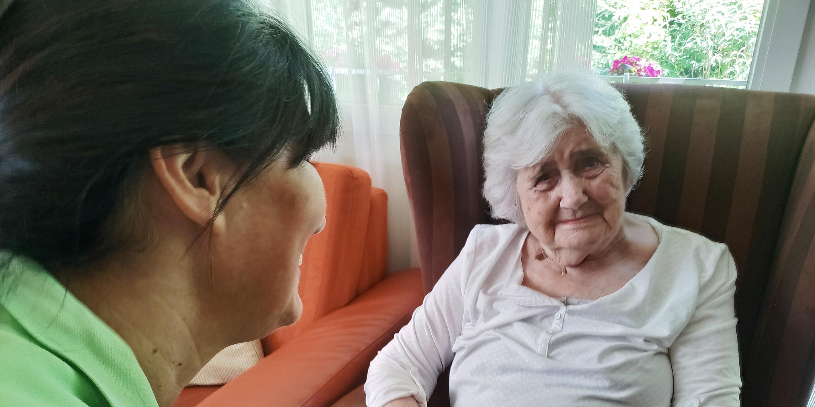 Eine alte weißhaarige Frau sitzt in einem roten Lehnsessel. Ein Pflegerin mit grünem Poloshirt hält ihre Hand.