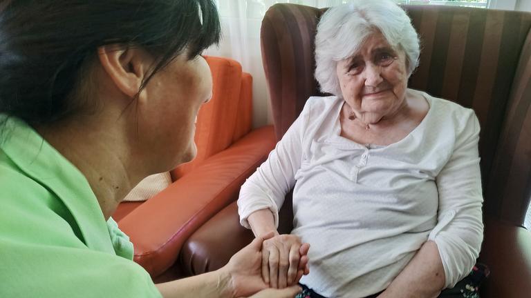 Eine alte weißhaarige Frau sitzt in einem roten Lehnsessel. Ein Pflegerin mit grünem Poloshirt hält ihre Hand.