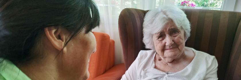 Eine alte weißhaarige Frau sitzt in einem roten Lehnsessel. Ein Pflegerin mit grünem Poloshirt hält ihre Hand.