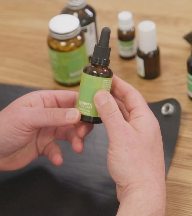 Person hält Flasche mit Bitterstoff-Tropfen in der Hand. Auf dem Tisch stehen verschiedene Produkte mit Bitterstoffen.