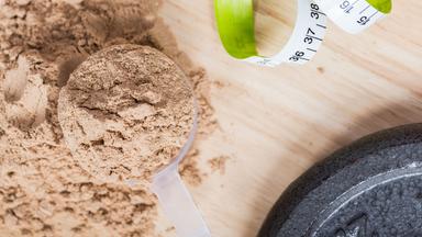 Ein Messbecher mit Molkenproteinpulver liegt auf einem Holztisch neben einem Maßband und einem Gewicht.