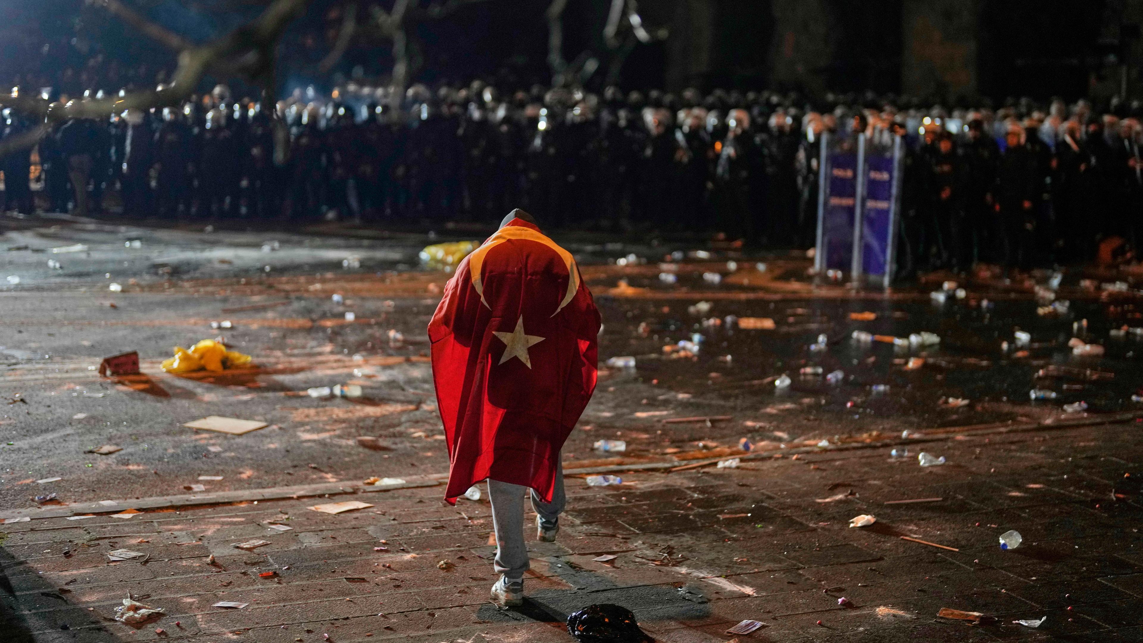 Ein Mann mit der türkischen Flagge auf dem Rücken steht während der Zusammenstöße bei einer Kundgebung gegen die Verhaftung von Istanbuls Bürgermeister Ekrem Imamoglu in Istanbul, Türkei, vor Polizisten, die gegen Unruhen vorgehen.Ein Mann mit der türkischen Flagge auf dem Rücken steht während der Zusammenstöße bei einer Kundgebung gegen die Verhaftung von Istanbuls Bürgermeister Ekrem Imamoglu in Istanbul, Türkei, vor Polizisten, die gegen Unruhen vorgehen.