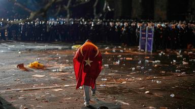 Ein Mann mit der türkischen Flagge auf dem Rücken steht während der Zusammenstöße bei einer Kundgebung gegen die Verhaftung von Istanbuls Bürgermeister Ekrem Imamoglu in Istanbul, Türkei, vor Polizisten, die gegen Unruhen vorgehen.Ein Mann mit der türkischen Flagge auf dem Rücken steht während der Zusammenstöße bei einer Kundgebung gegen die Verhaftung von Istanbuls Bürgermeister Ekrem Imamoglu in Istanbul, Türkei, vor Polizisten, die gegen Unruhen vorgehen.