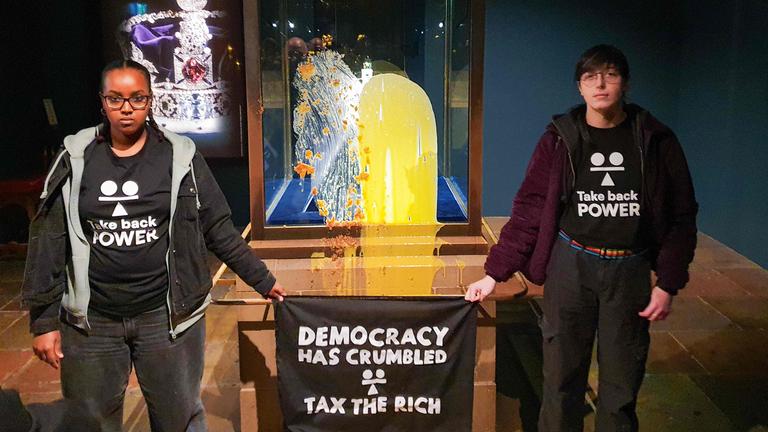 Ein Foto vom 06.12.2025 zeigt Anhänger der zivilen Widerstandsgruppe "Take Back Power" mit einem Banner mit der Aufschrift "Democracy has crumbled – Tax The Rich", nachdem sie Dessert und Vanillesoße über die Vitrine mit den Kronjuwelen im Tower of London geschüttet hatten.
