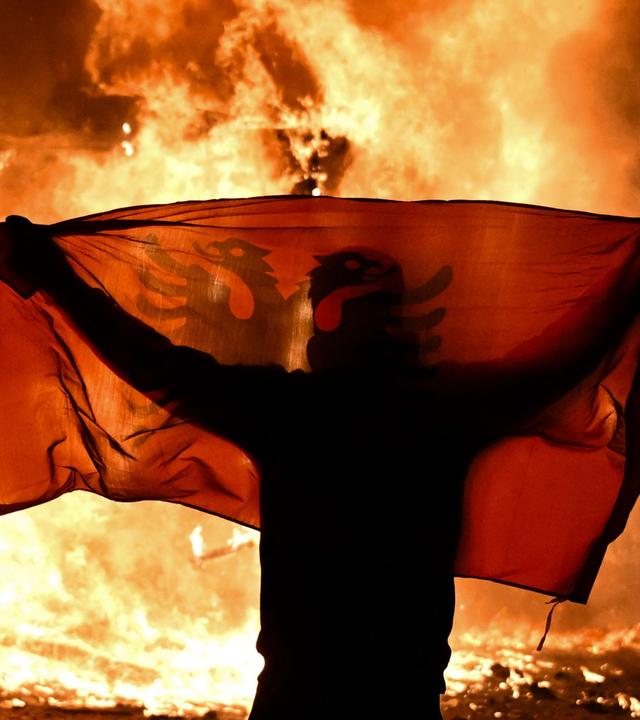 Ein Protestant hält am15.10.2026 während der Proteste gegen die Regierung in Tirana vor einem bennenden Kiosk die albanische Flagge hoch.