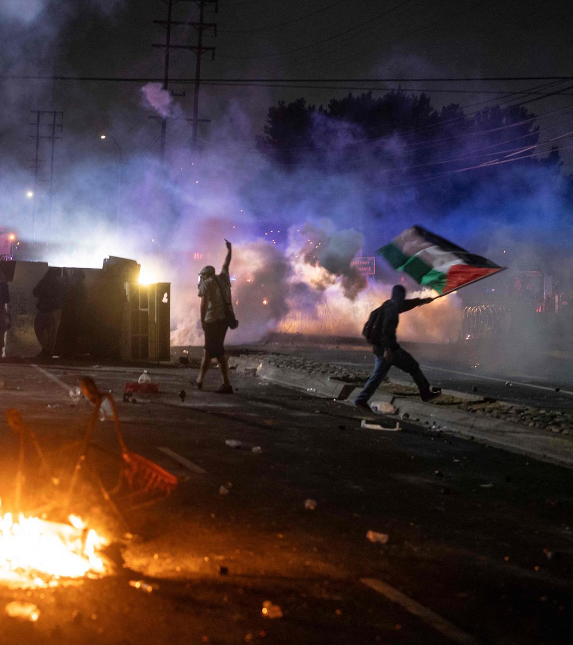 USA, Los Angeles: Protestierende stehen in der Nacht auf einer Straße. Im Vordergrund brennt etwas, ein Demonstrierender (r) hält eine palästinensische Flagge, im Hintergrund stehen vom Rauch verdeckte Autos.