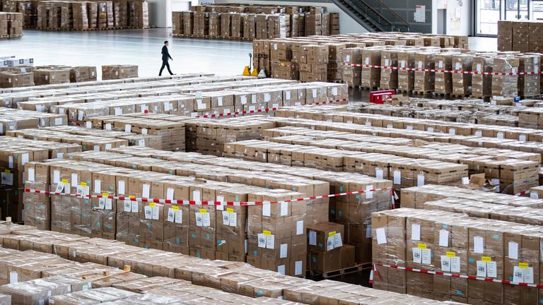 Ein Mann mit Schutzmaske geht an zahlreichen Kartons mit Schutzausrüstung in Millionenzahl vorbei.